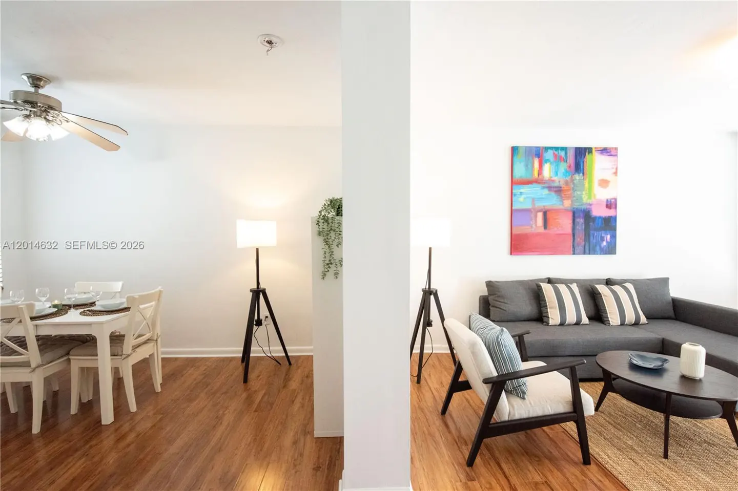 A bright, modern living space with a dining area, gray sofa, and colorful artwork. The floors are wood and there are tripod lamps.