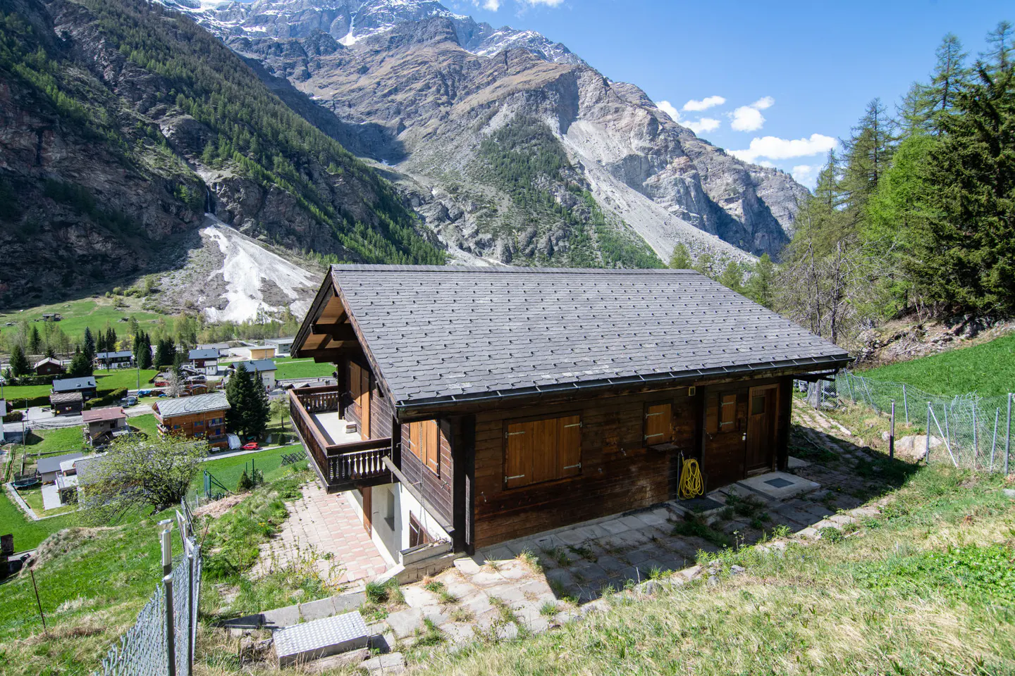 A brown chalet with a gray roof sits on a grassy hill with mountains in the background.
