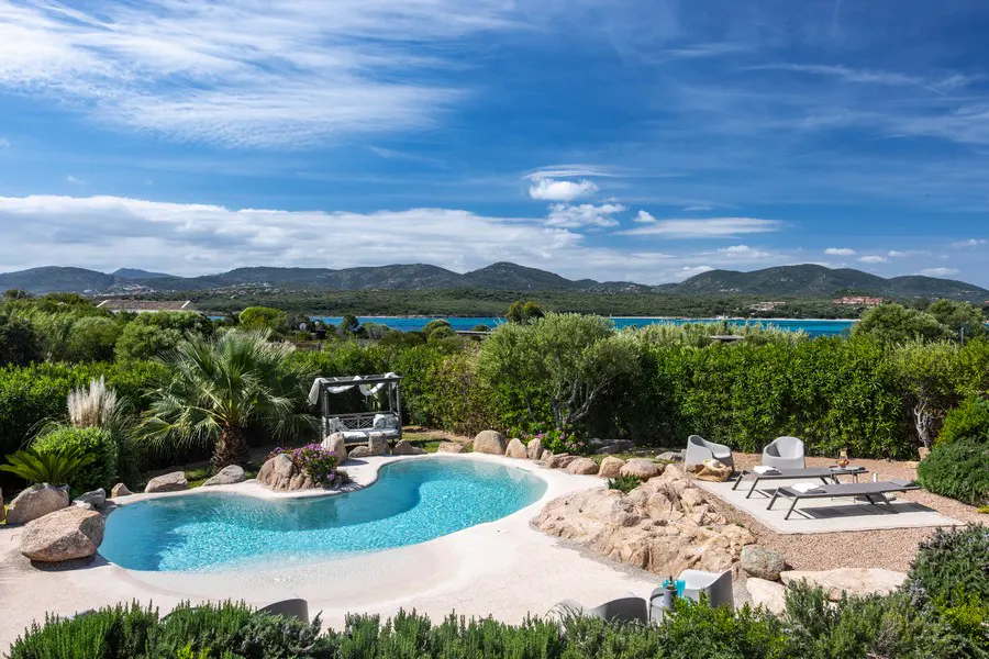 Scenic view of a kidney-shaped pool with lounge chairs, a cabana, and a view of the ocean and mountains.