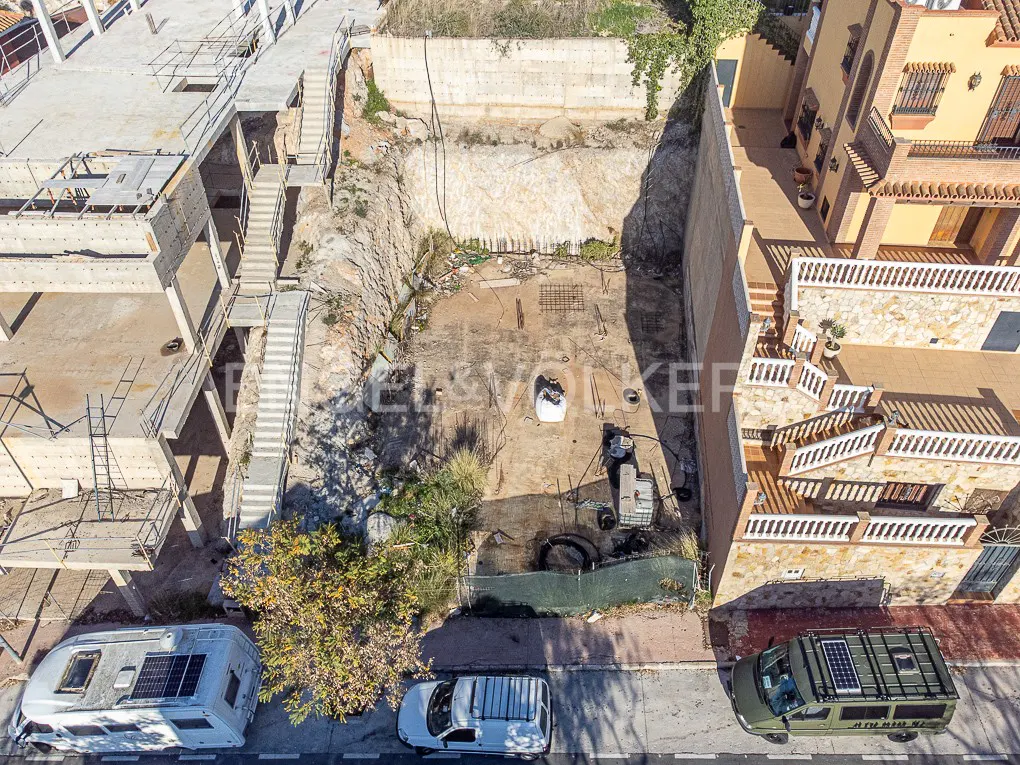 Aerial view of a vacant lot between buildings, with parked vehicles on the street. The lot has a retaining wall and some construction materials.