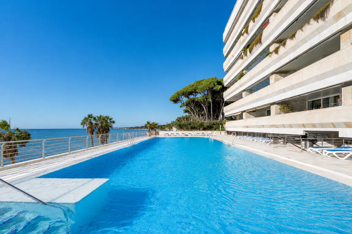 A bright blue swimming pool overlooking the ocean, next to a modern multi-story apartment building with balconies.