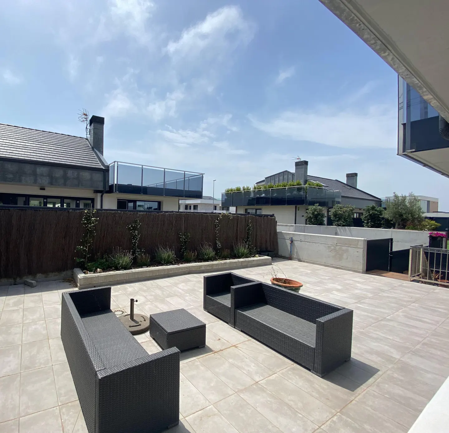 Outdoor patio with gray wicker furniture set, including sofas and a table, on a tiled floor. A fence and buildings are in the background under a blue sky.