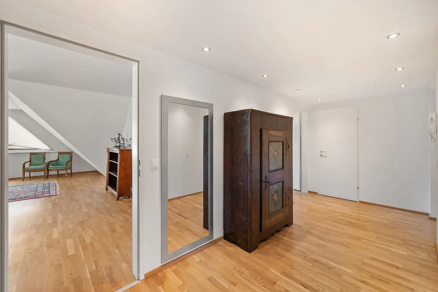 Hallway with wood floors, white walls, and recessed lighting. A large, dark wood cabinet and a full-length mirror are visible. An open doorway leads to another room.