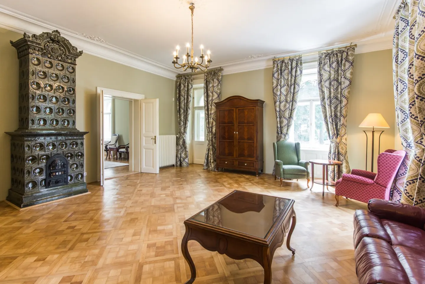 A spacious living room with parquet floors, a tiled fireplace, and patterned curtains. Furniture includes a brown wardrobe, chairs, and a coffee table.