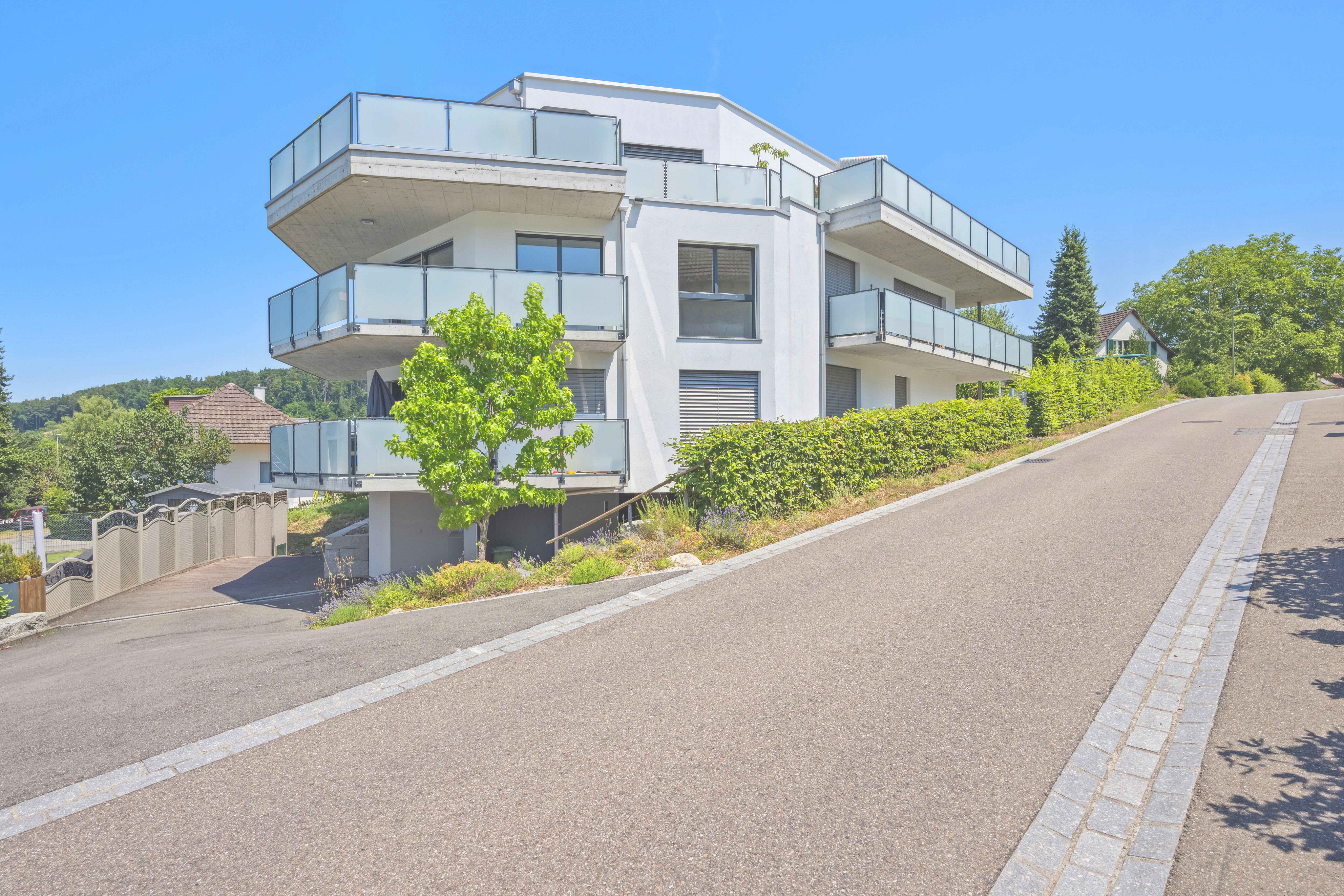 Modern white multi-story apartment building with glass balconies, situated on a sloped street under a clear blue sky. [AI-generated text]