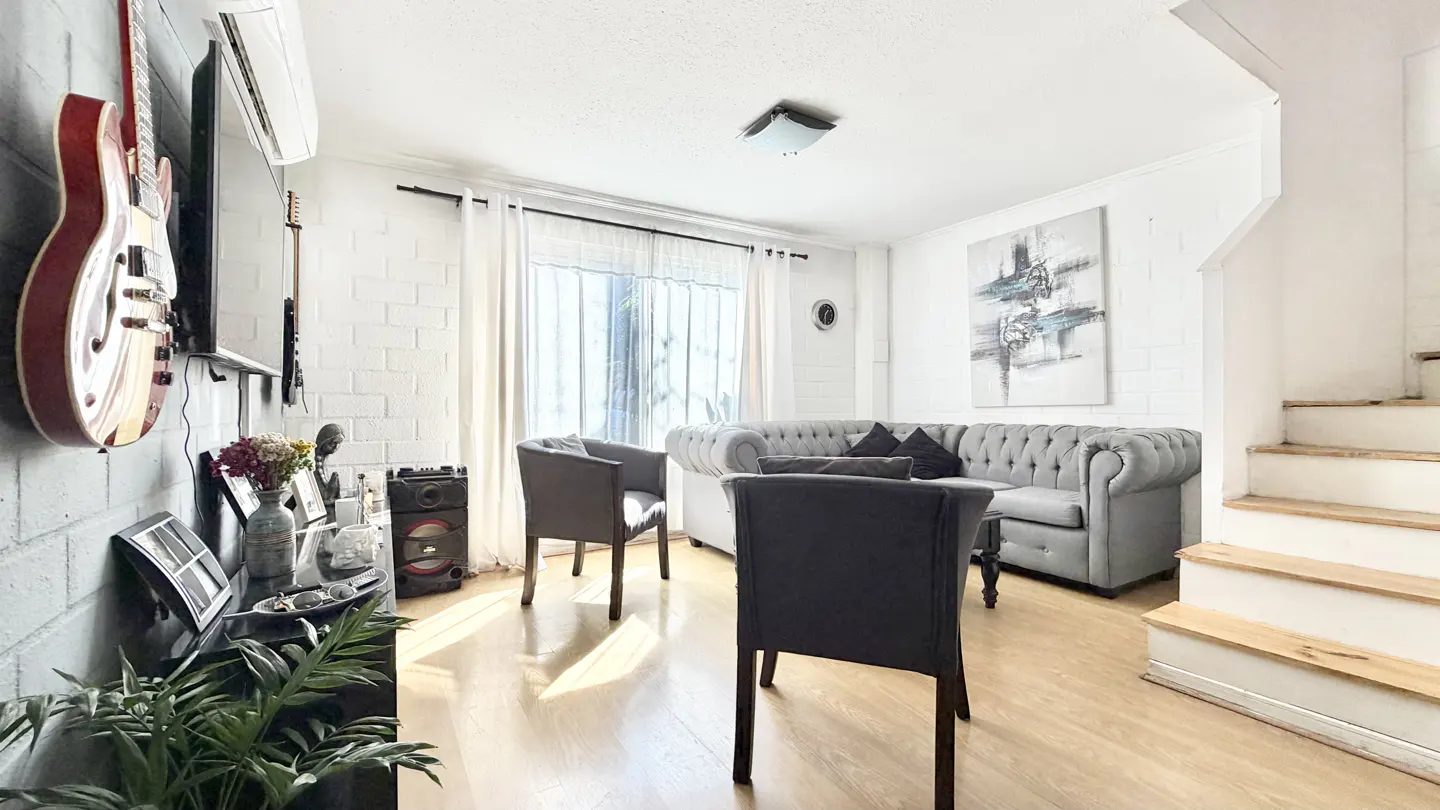 Bright living room with hardwood floors, gray sofa, two chairs, and guitars hanging on a white brick wall. Stairs are visible on the right.