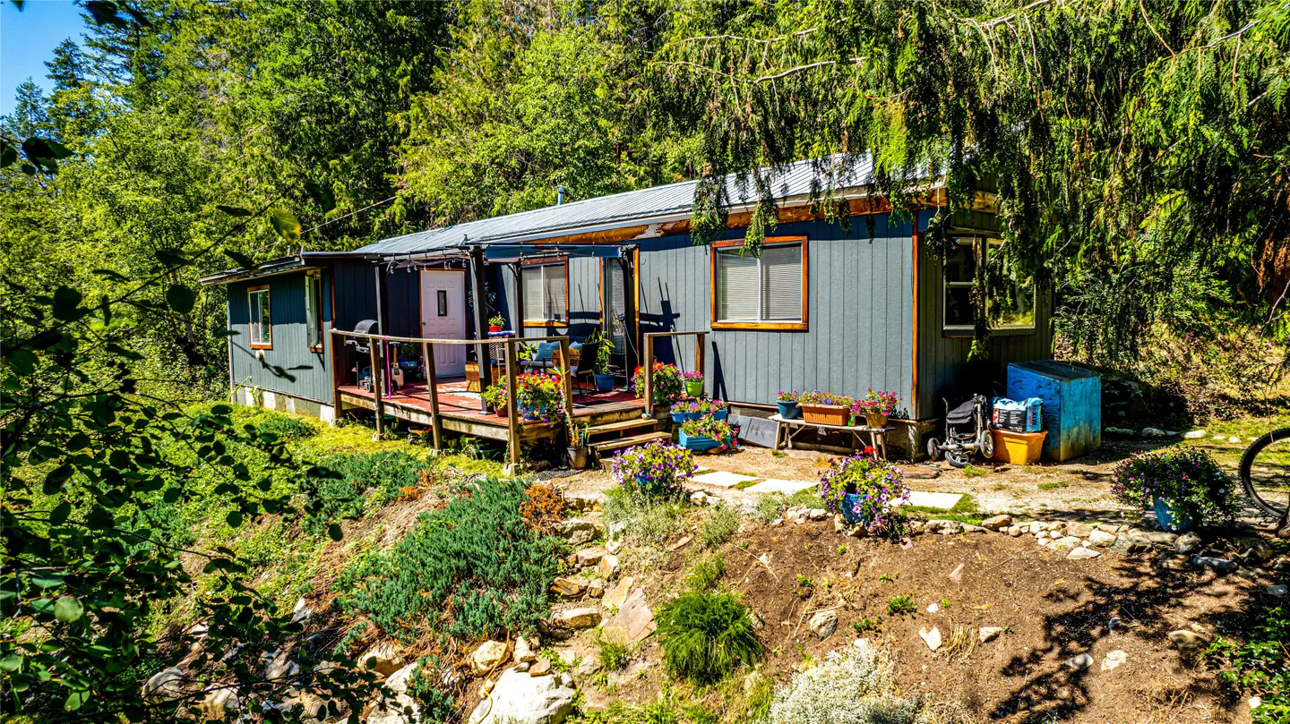 Exterior view of a gray house with a wooden porch, surrounded by lush greenery and trees. Colorful flowers adorn the porch and garden.
