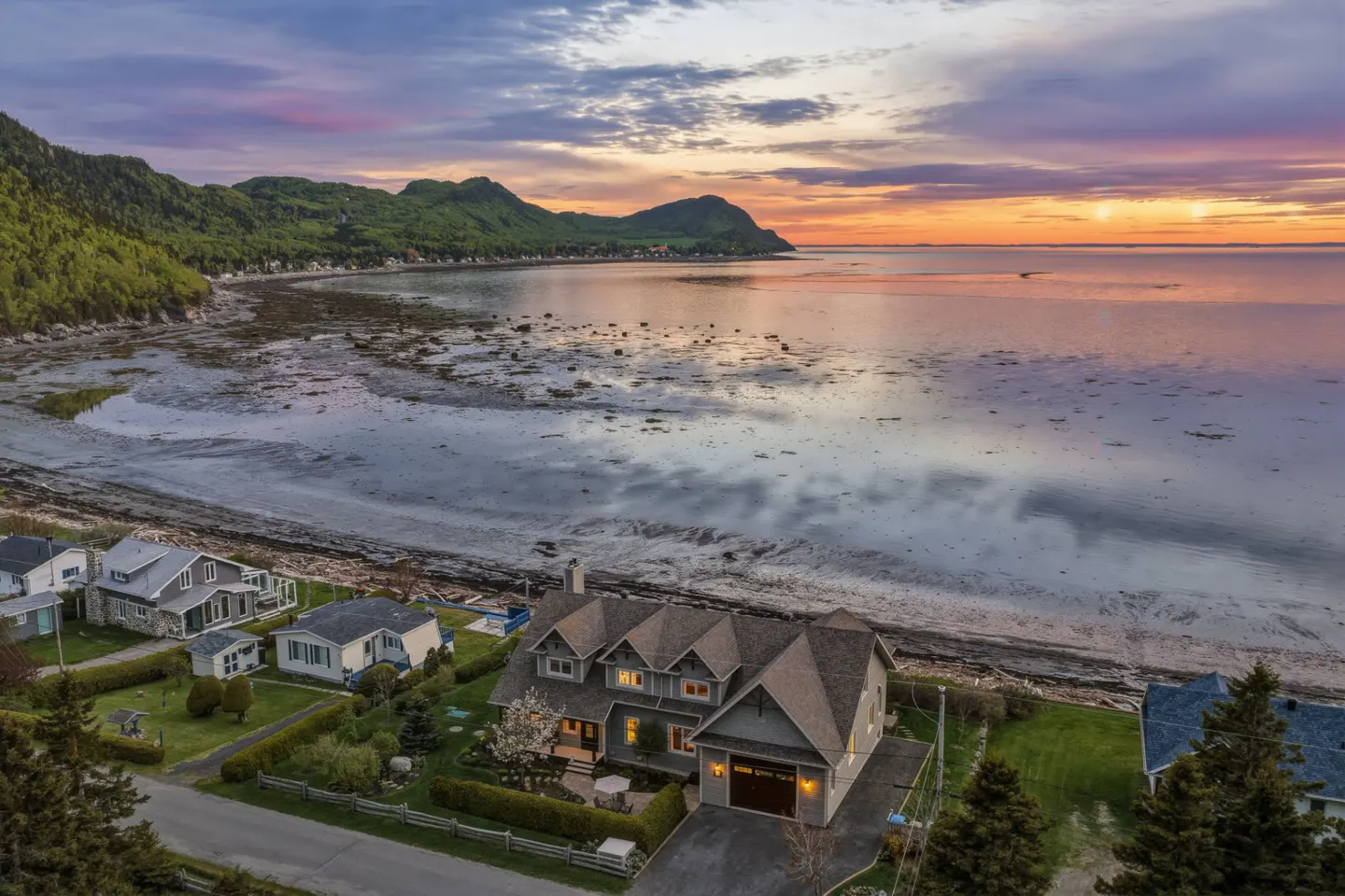 Aerial view of a coastal home at sunset, featuring a calm bay, lush green mountains, and a vibrant orange-purple sky.