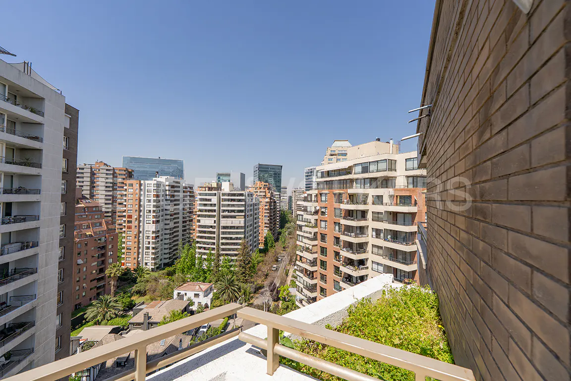 Cityscape view from a balcony. Tall buildings, trees, and a clear blue sky are visible. A brick wall is on the right.