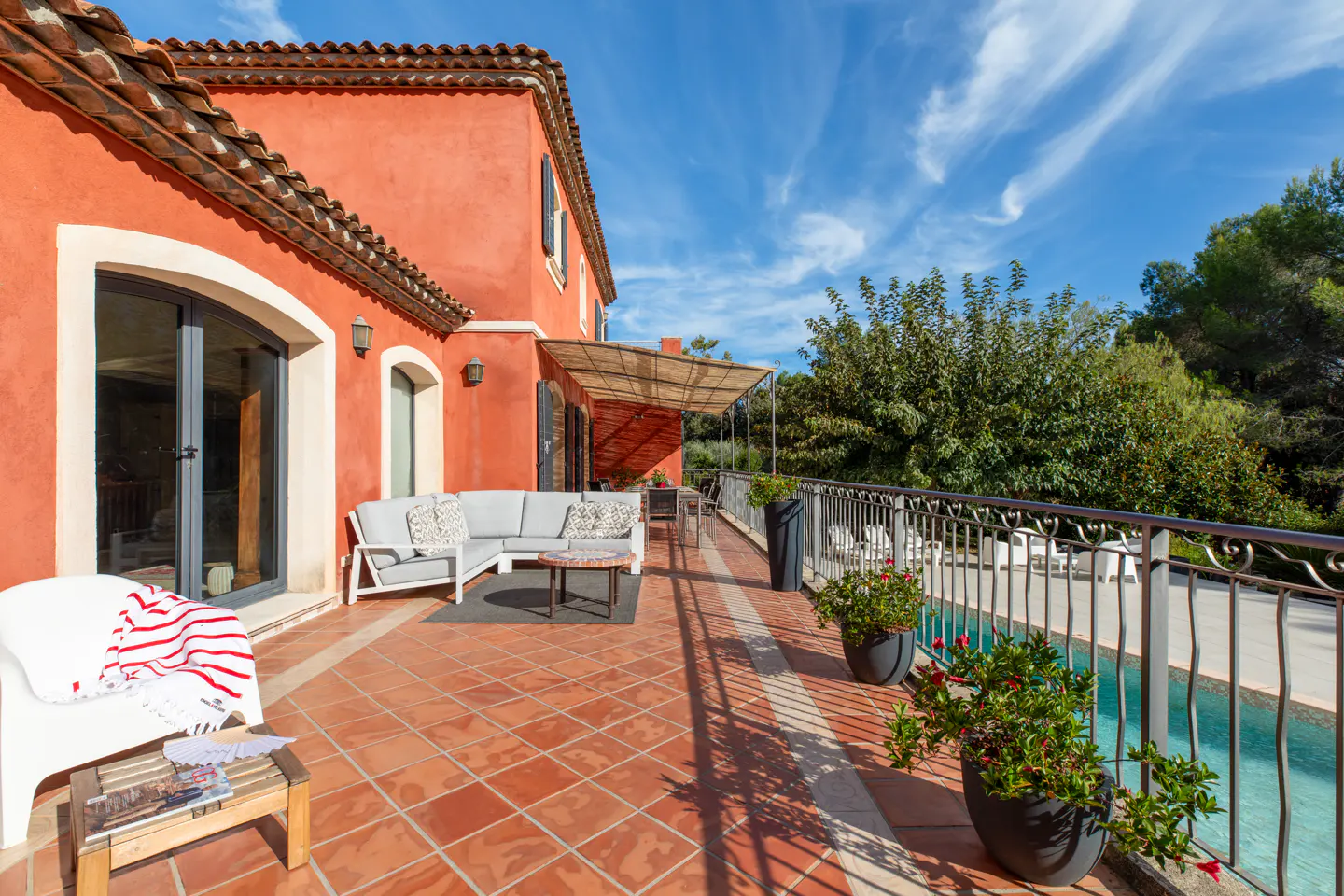 Terracotta patio with gray sofa, table, and potted plants outside a red stucco house with a blue sky.