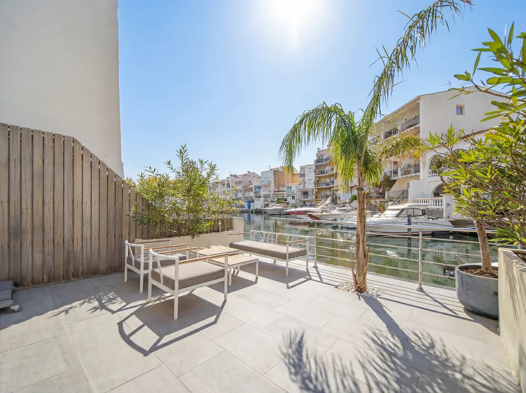 Outdoor patio with white furniture overlooking a canal with boats, palm trees, and white buildings under a bright blue sky.