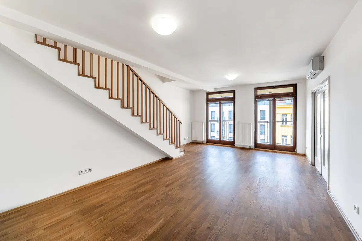 Bright, empty room with hardwood floors, white walls, and a staircase with a wooden railing. Balcony doors let in natural light.
