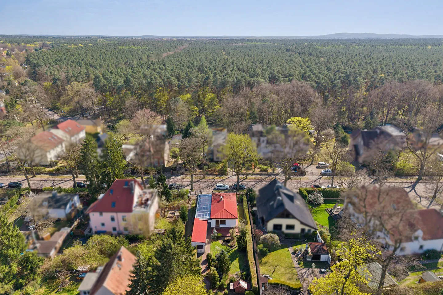 Aerial view of a suburban neighborhood with houses, trees, and a large forest in the background. The houses have red and dark roofs.