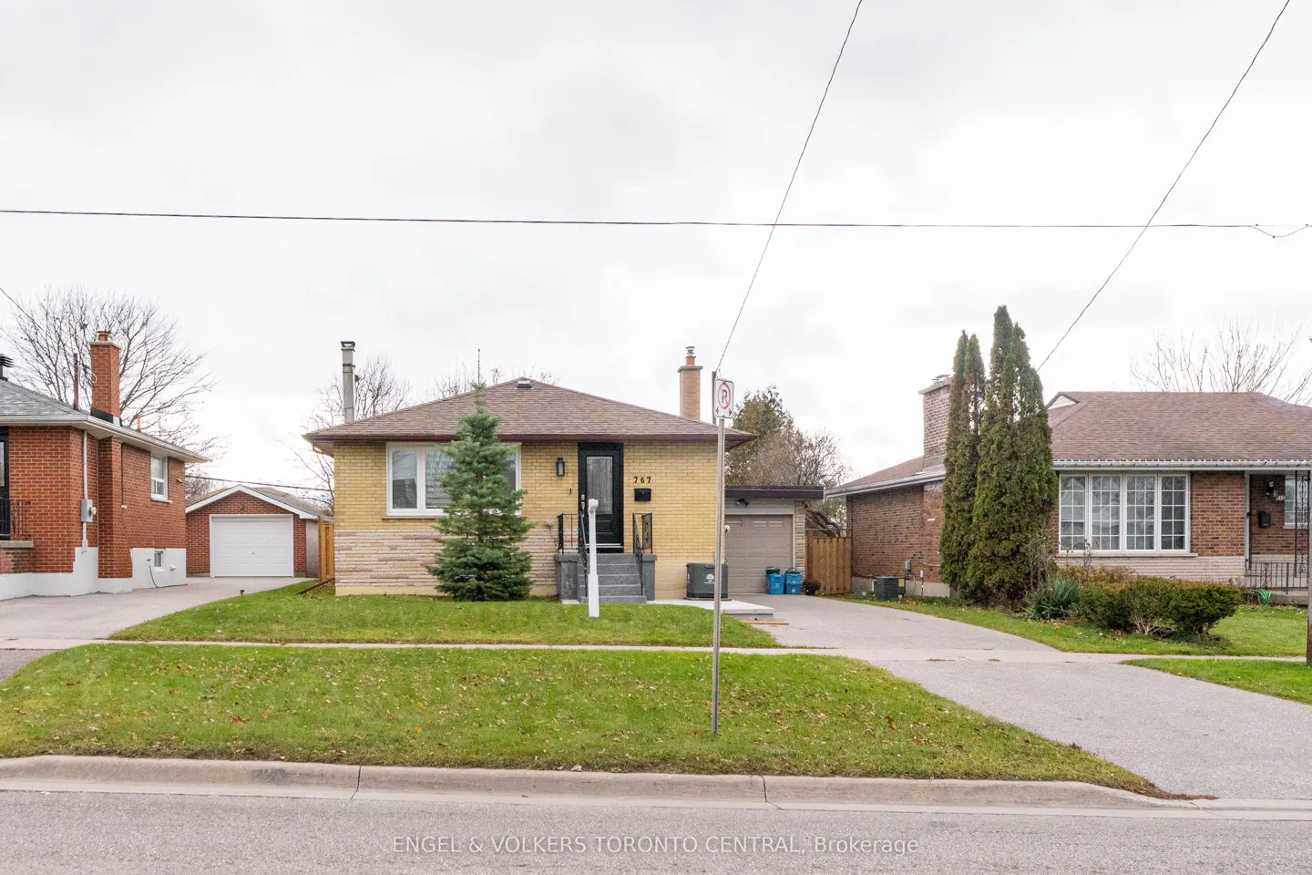 A yellow brick bungalow with a brown roof, green lawn, and a paved driveway on a cloudy day.