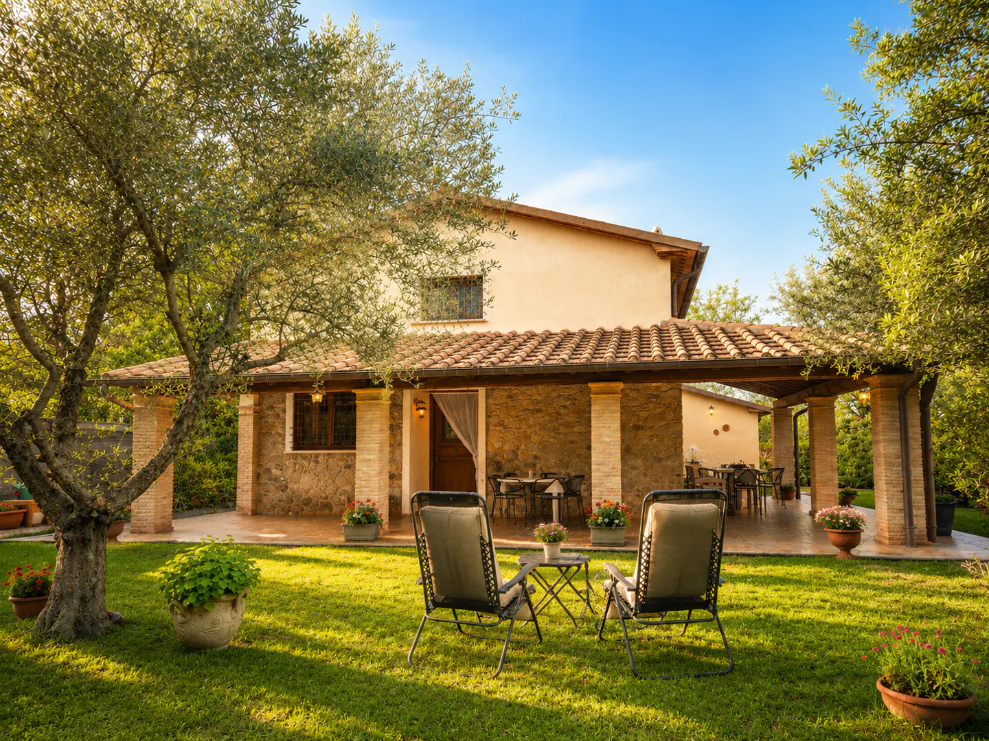 A tan two-story house with a red tile roof and a covered patio with chairs on the lawn.