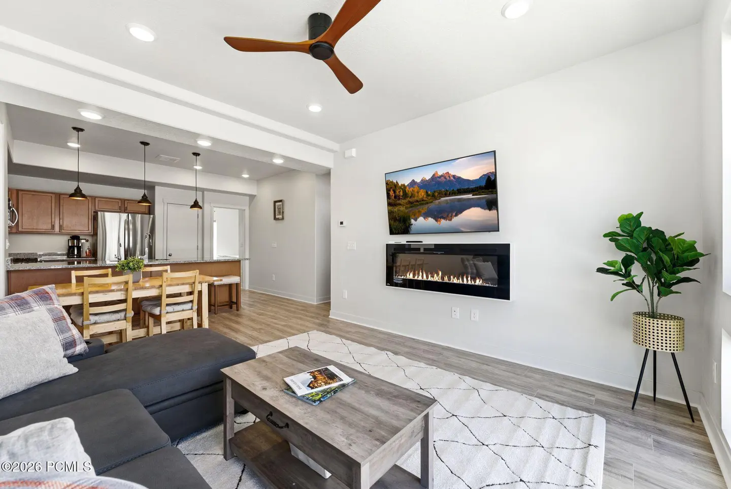 Living room with gray sectional, wood coffee table, TV above fireplace, and open kitchen with dining table.