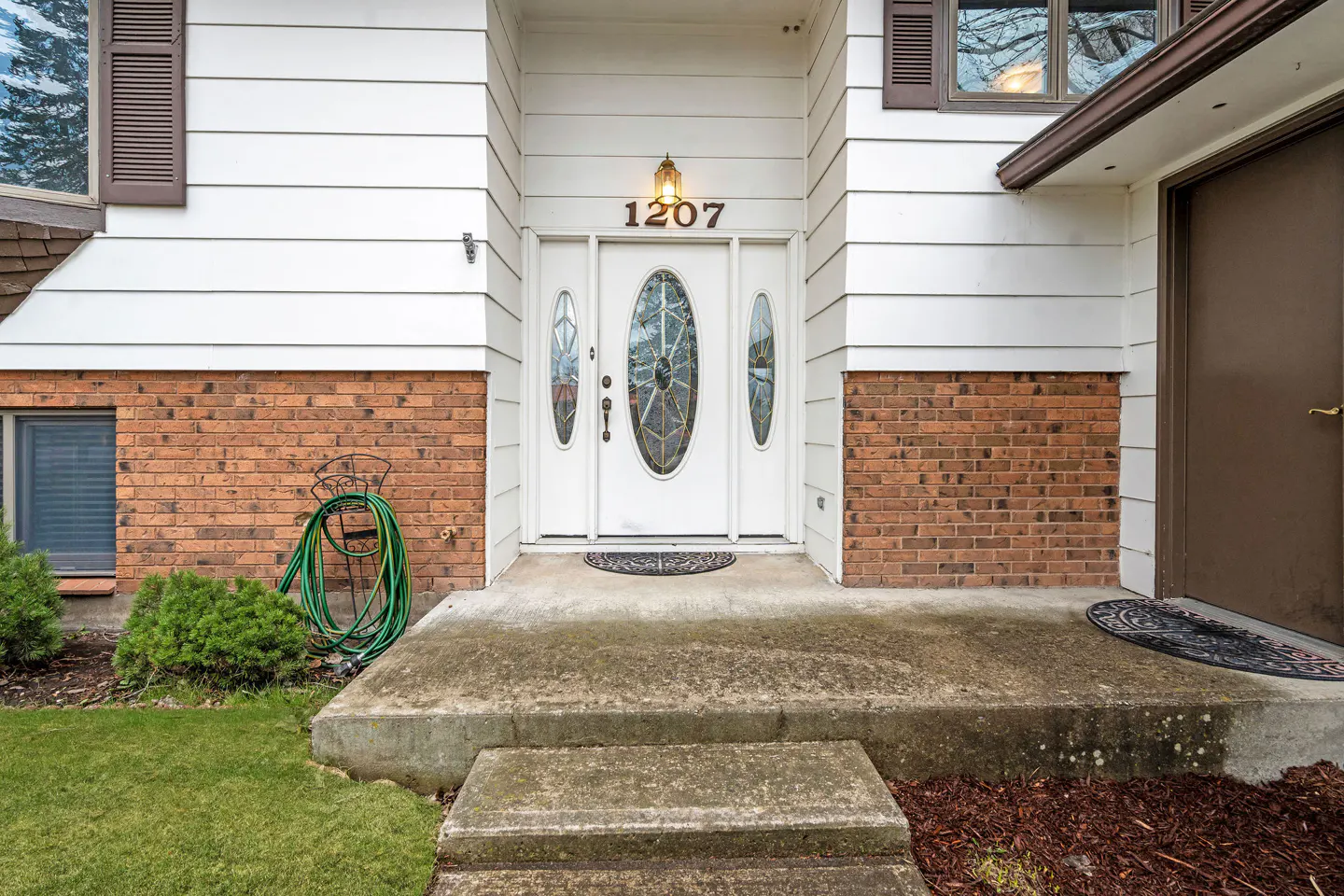 Exterior shot of a home's front entrance with a white door, brick accents, and green lawn. The house number "1207" is above the door.