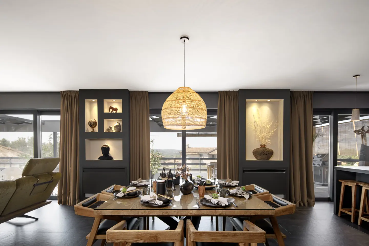 Dining room with a wood table, chairs, and a woven pendant light. Dark gray walls with recessed shelves and brown curtains frame the windows.