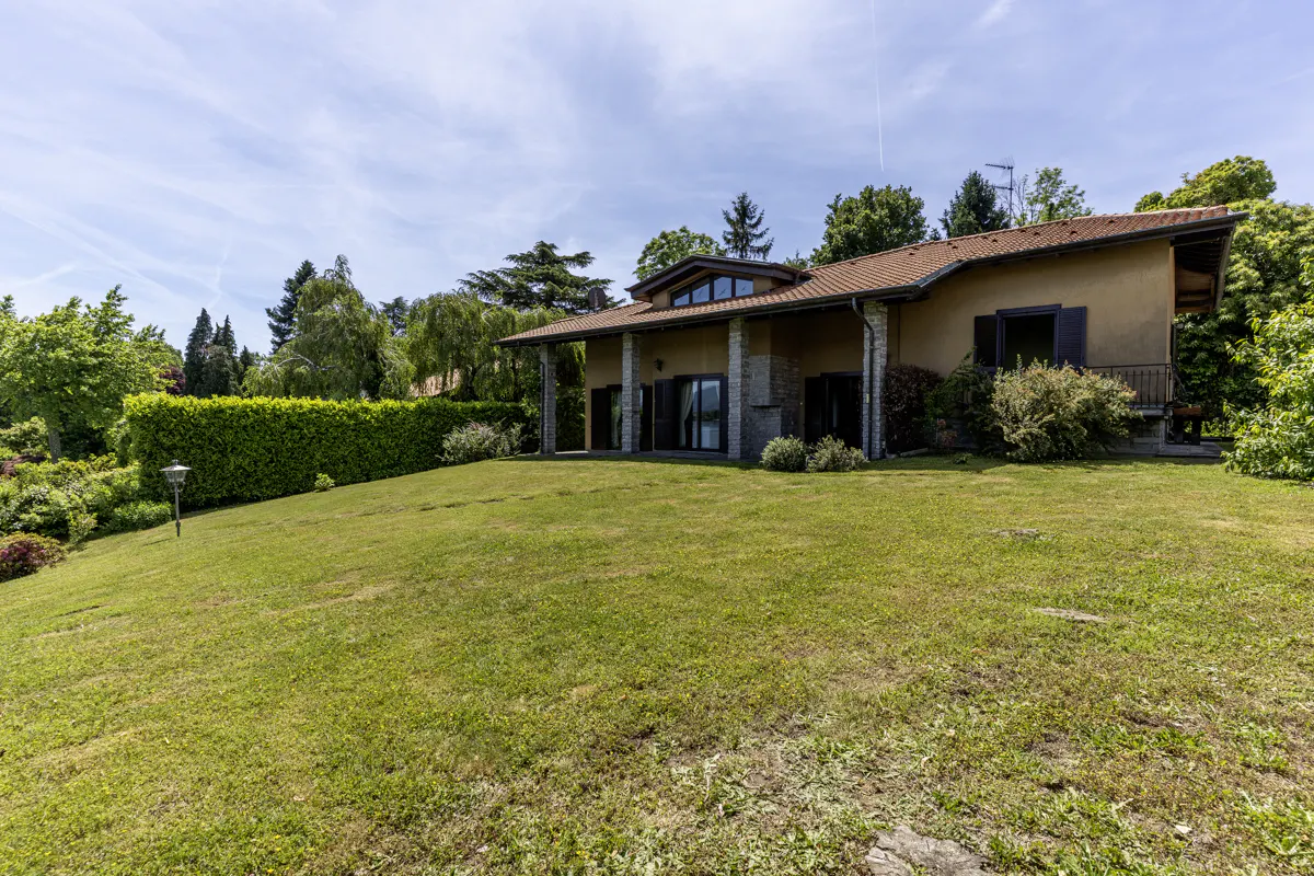A tan house with a red tile roof sits on a grassy hill under a blue sky with trees in the background.