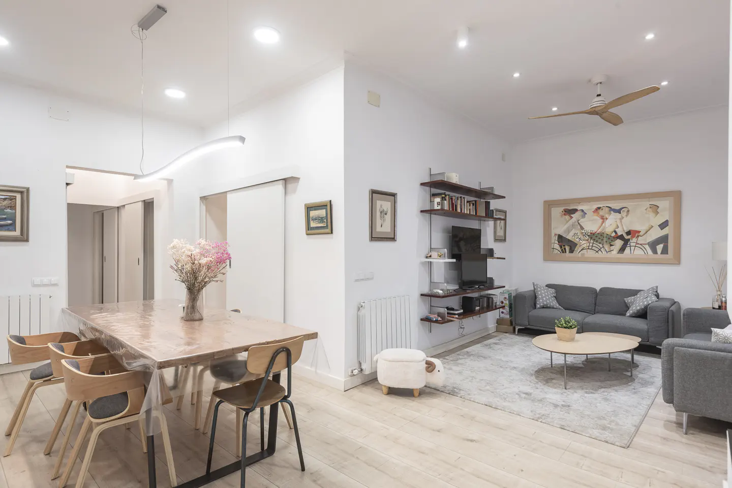 Modern open-plan living area with a wooden dining table, grey sofa, wall shelving, and light wood flooring.