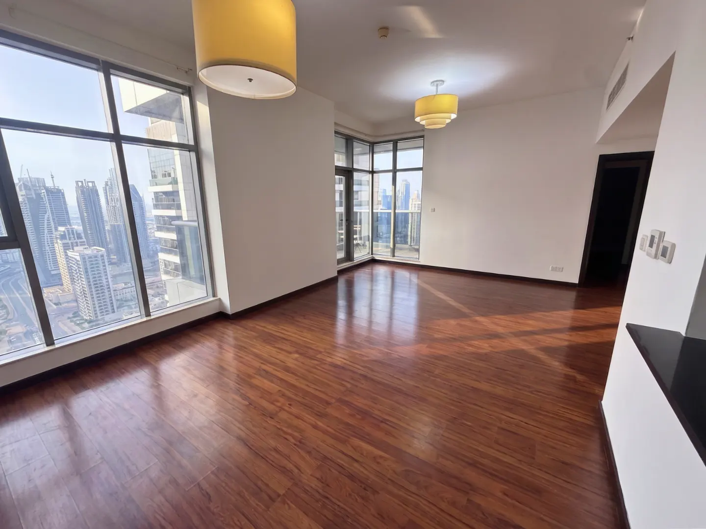 A bright, empty room with wood floors, white walls, and large windows showing a city skyline. Two yellow light fixtures hang from the ceiling.