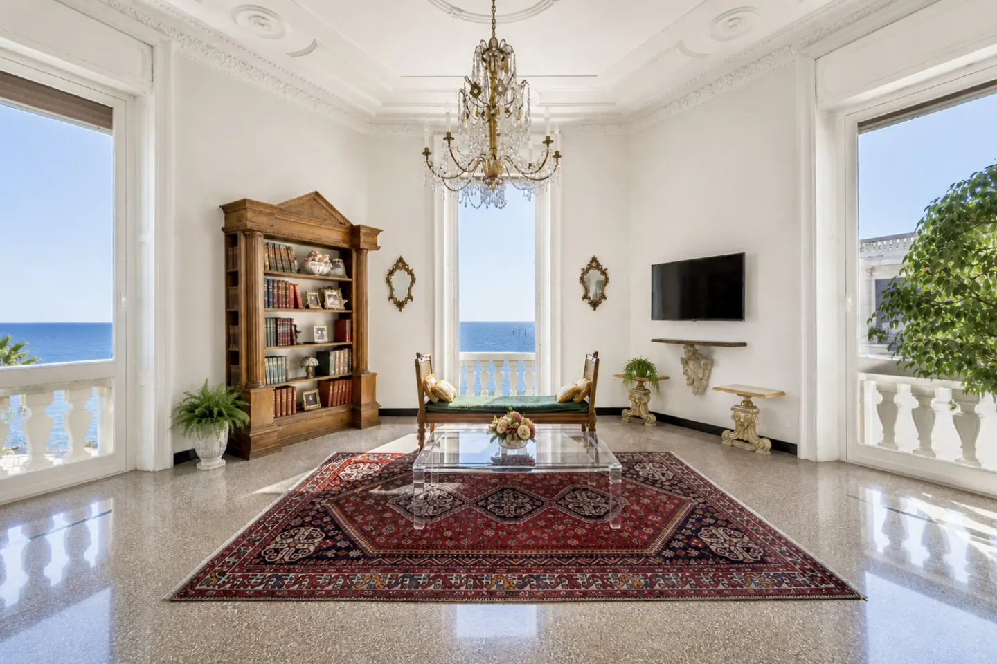 Bright living room with ocean view. Features a bookshelf, chandelier, rug, and balcony.