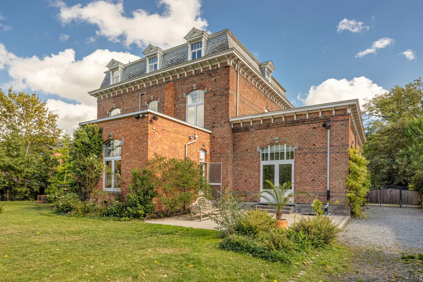 Exterior view of a large, red brick house with a gray roof, white trim, and green lawn under a blue sky with white clouds.