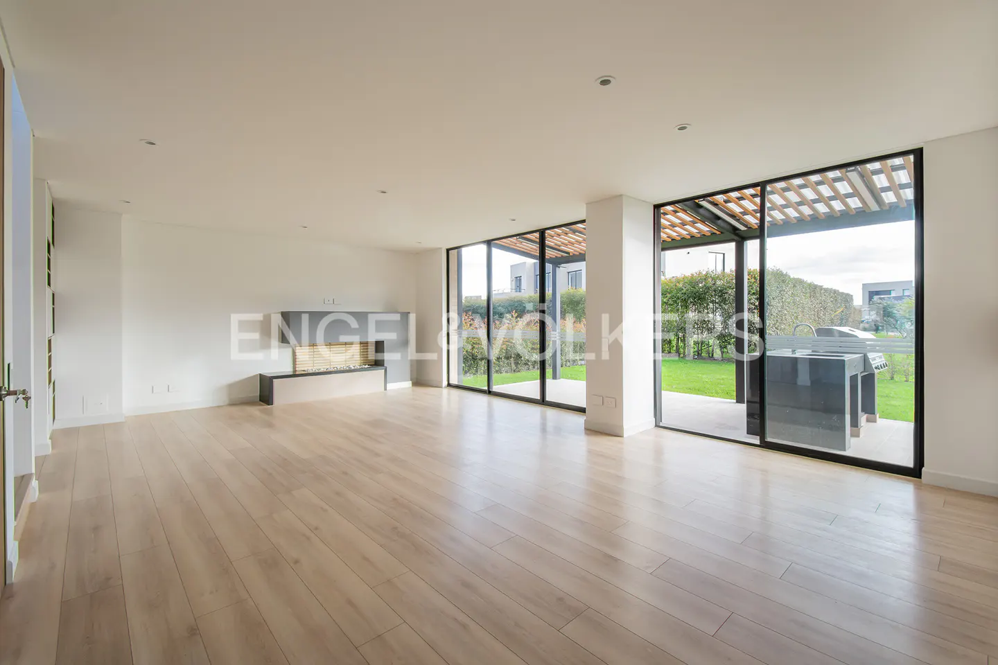 Bright, empty living room with light wood floors, a gray fireplace, and large windows overlooking a green lawn.