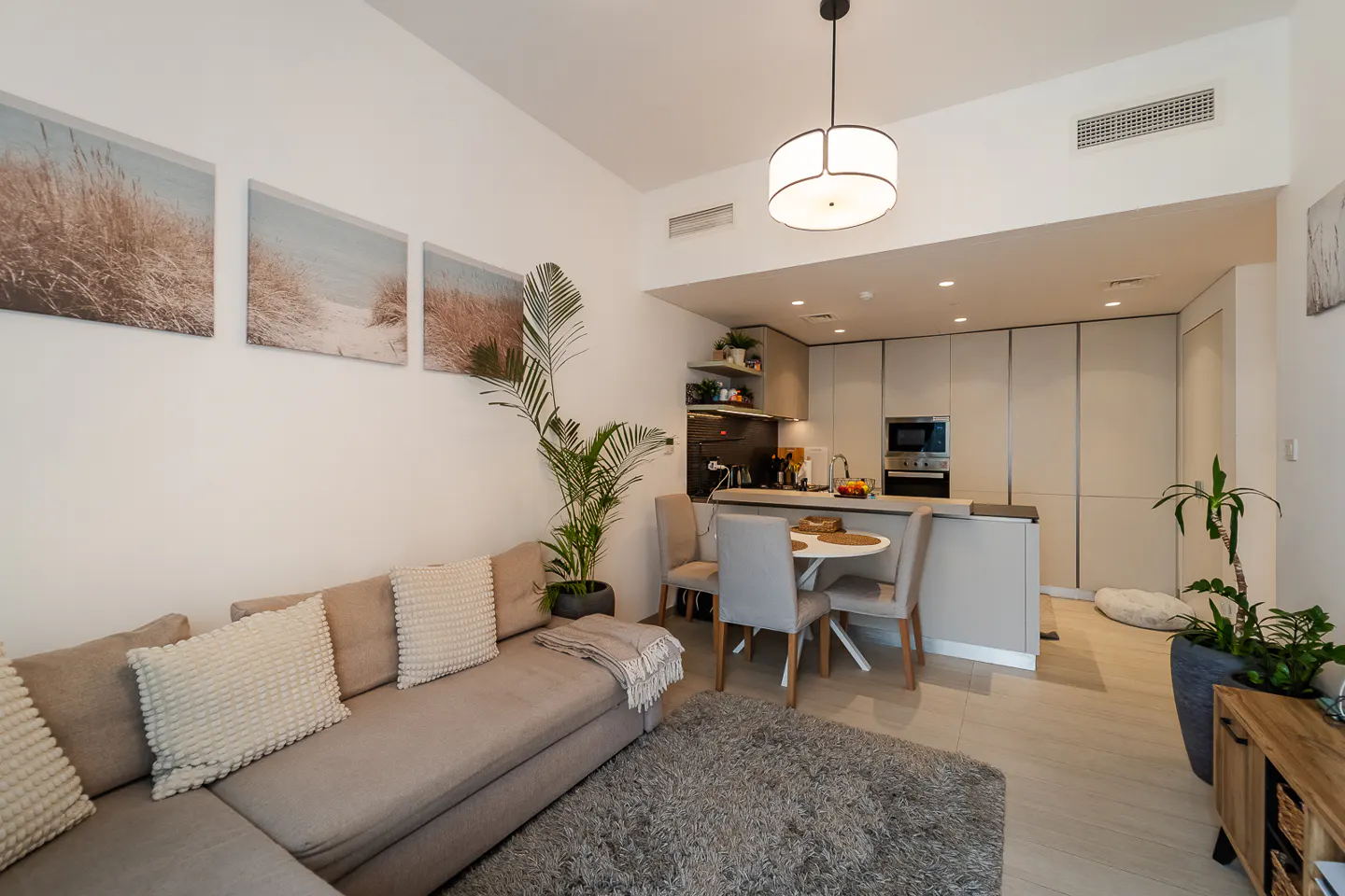 Living room with a beige sectional sofa, a round dining table with four chairs, and a modern kitchen in the background. The walls are decorated with beach-themed artwork.