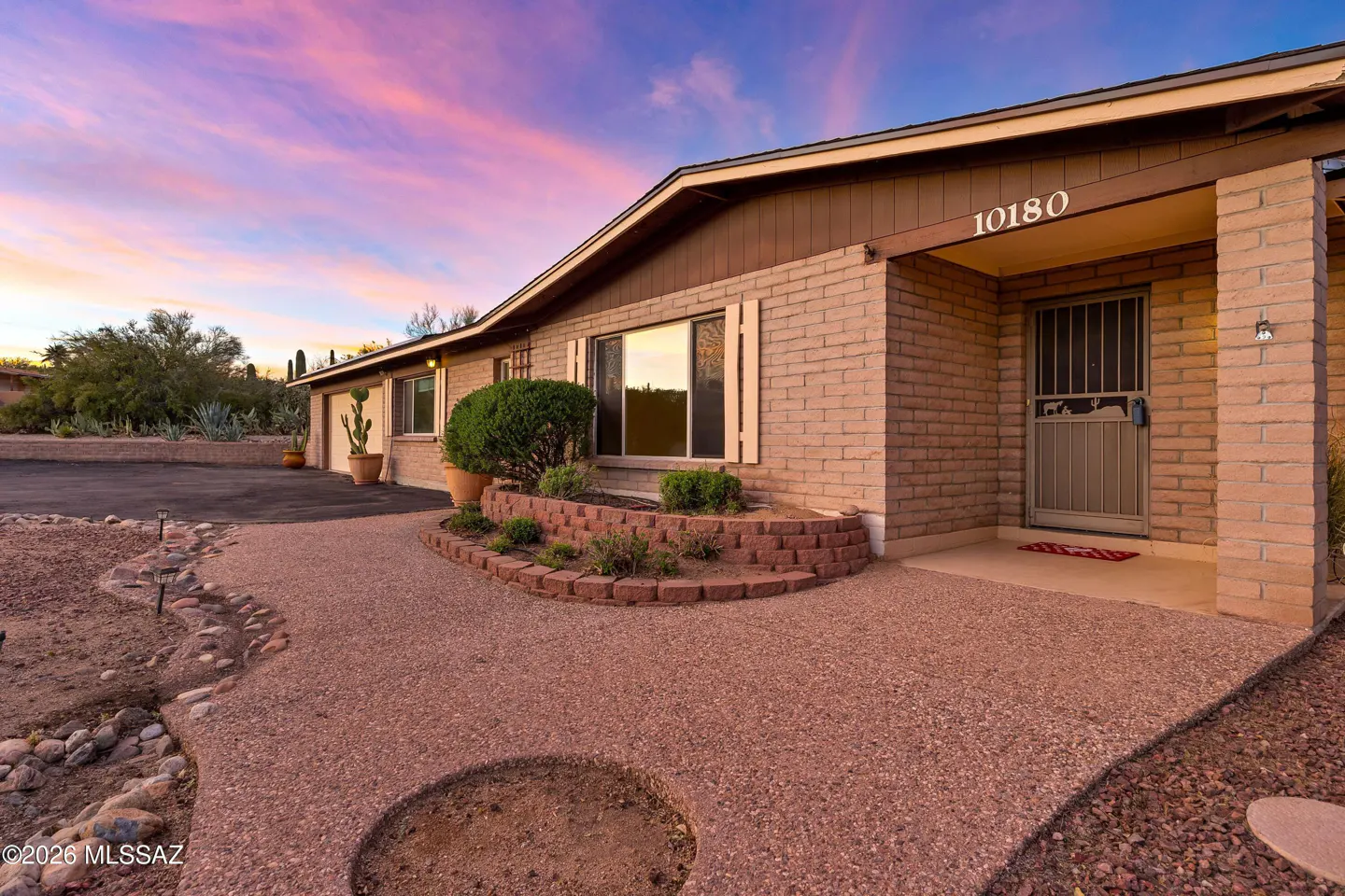 Exterior shot of a single-story brick house with brown trim under a pink and purple sky. A walkway leads to the front door.
