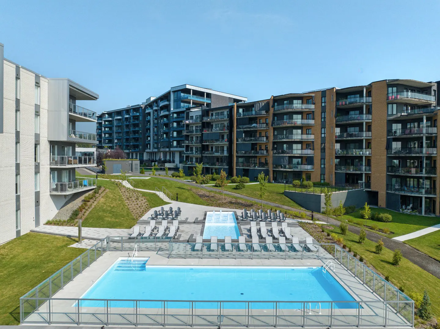 Apartment complex with a pool. The pool is surrounded by lounge chairs and green grass. Balconies are visible on the apartments.