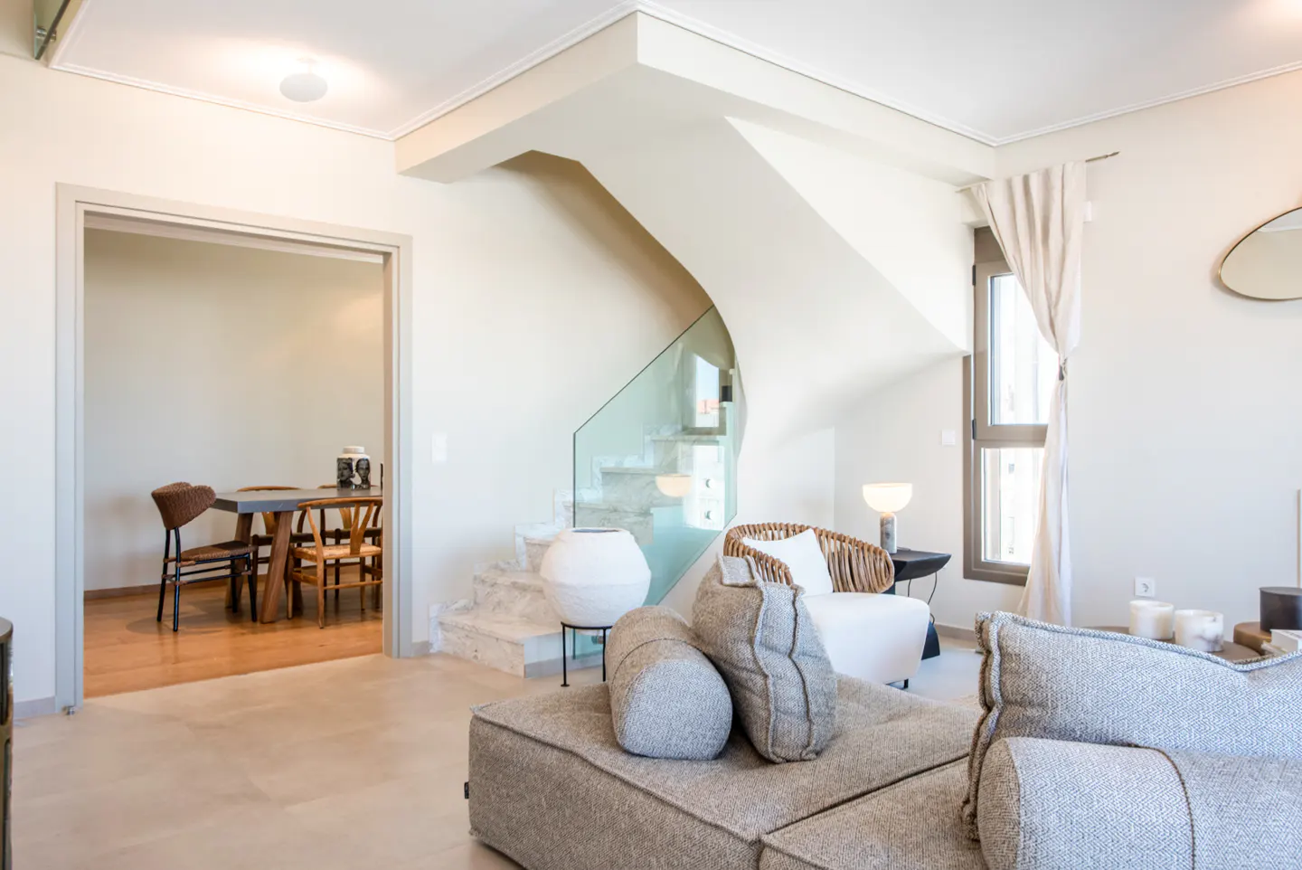 Bright living room with a gray sectional sofa, a winding staircase with glass railing, and a dining room visible through an open doorway.