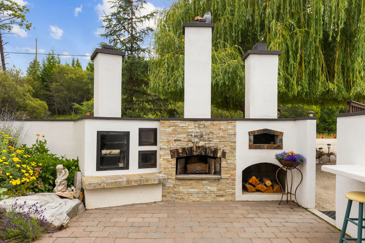 Outdoor stone fireplace and pizza oven with white stucco chimneys, wood storage, and garden seating on a paved patio.