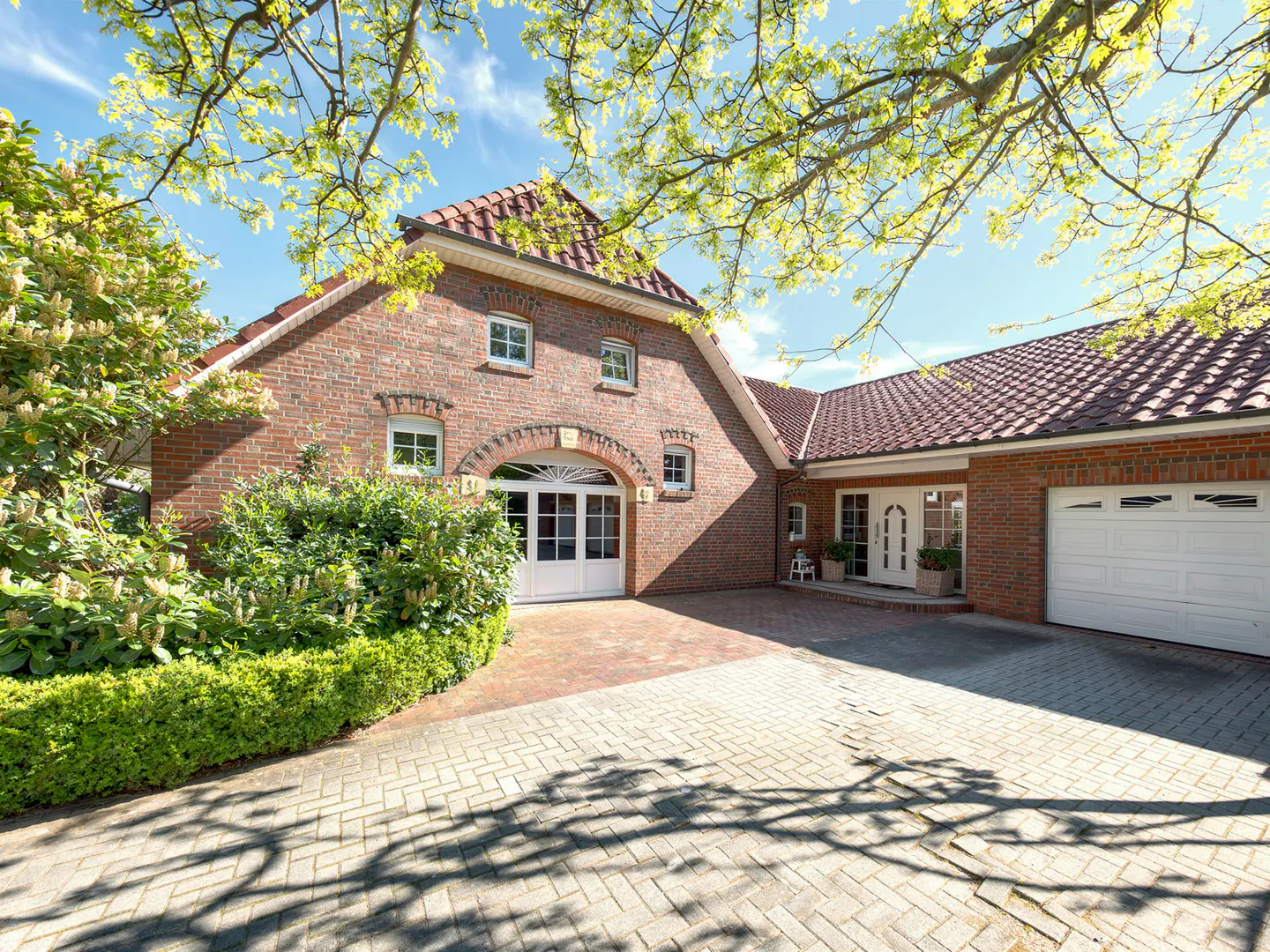 Exterior of a red brick house with a red tile roof, white trim, and a brick driveway. Green trees frame the house.