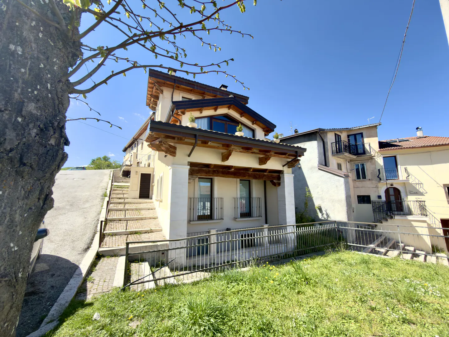 Exterior view of a two-story house with a brown roof, cream walls, and a metal fence, set on a grassy hill under a blue sky.