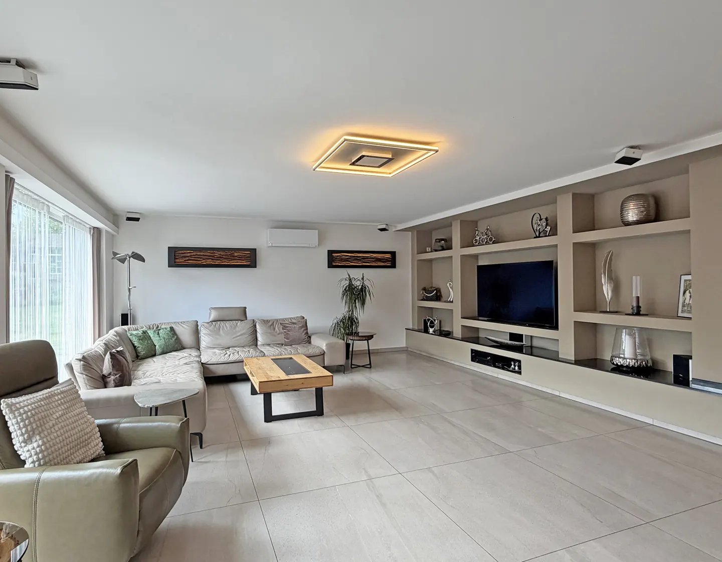 Modern living room with a beige sectional sofa, leather armchair, wooden coffee table, and built-in wall shelving with a TV.