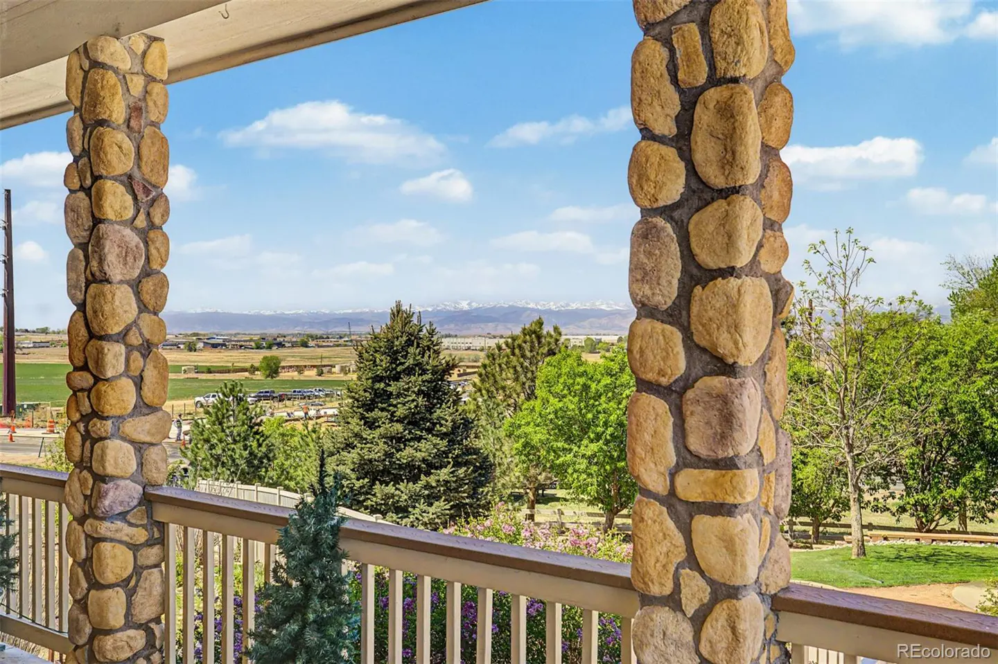 View from a porch with stone pillars, a wooden railing, and a scenic view of trees, fields, and distant mountains under a blue sky.