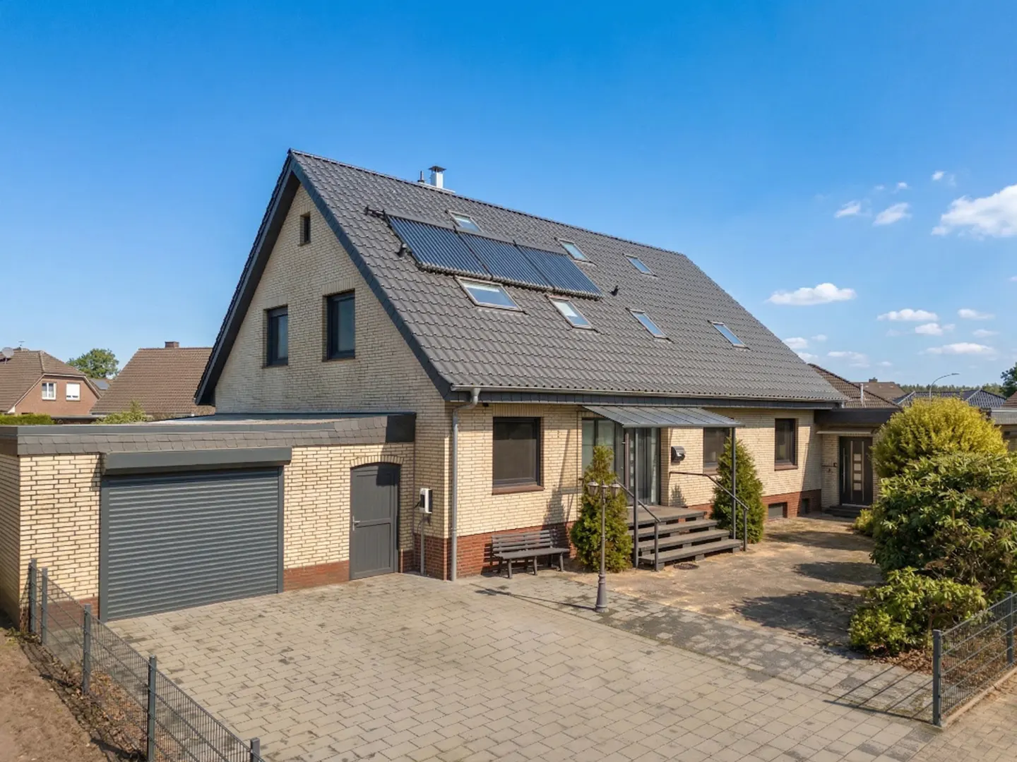 Two-story brick house with a gray roof, solar panels, and a detached garage on a paved driveway under a blue sky.