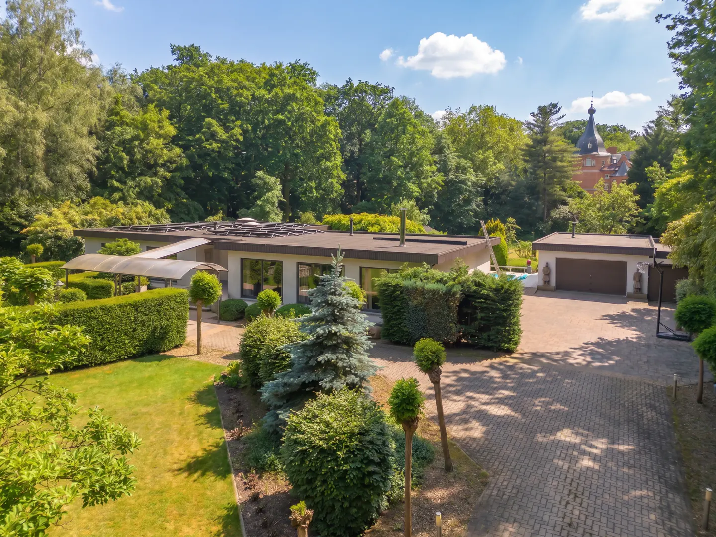 Modern flat-roofed house with a paved driveway, lush green landscaping, and a detached garage on a sunny day.