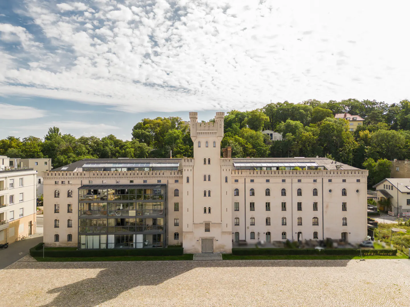 Exterior of a large, light-beige building with a central tower, glass facade, and green trees in the background.