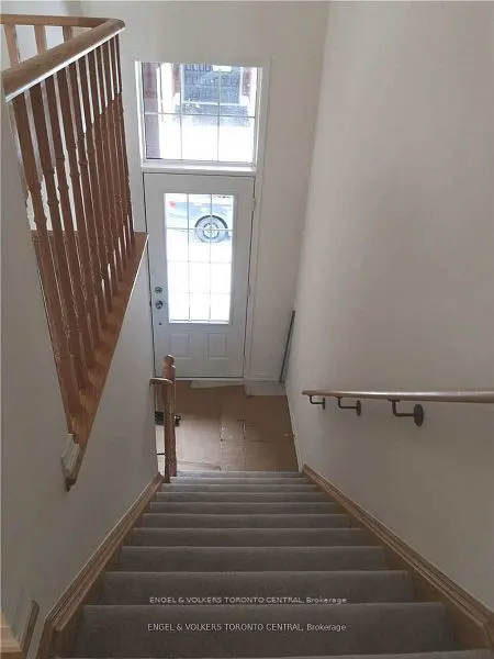 Interior view of carpeted stairs leading to a white front door with a window above. Wooden banister on the left, handrail on the right. Cream-colored walls.