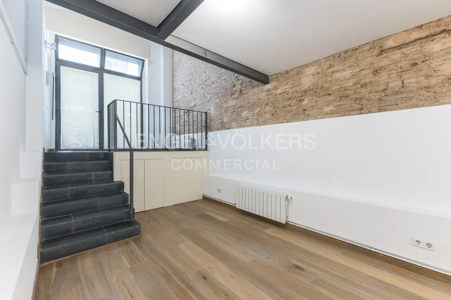 Interior view of a commercial space with wooden floors, brick wall, stairs, and Engel & Volkers logo on the wall.