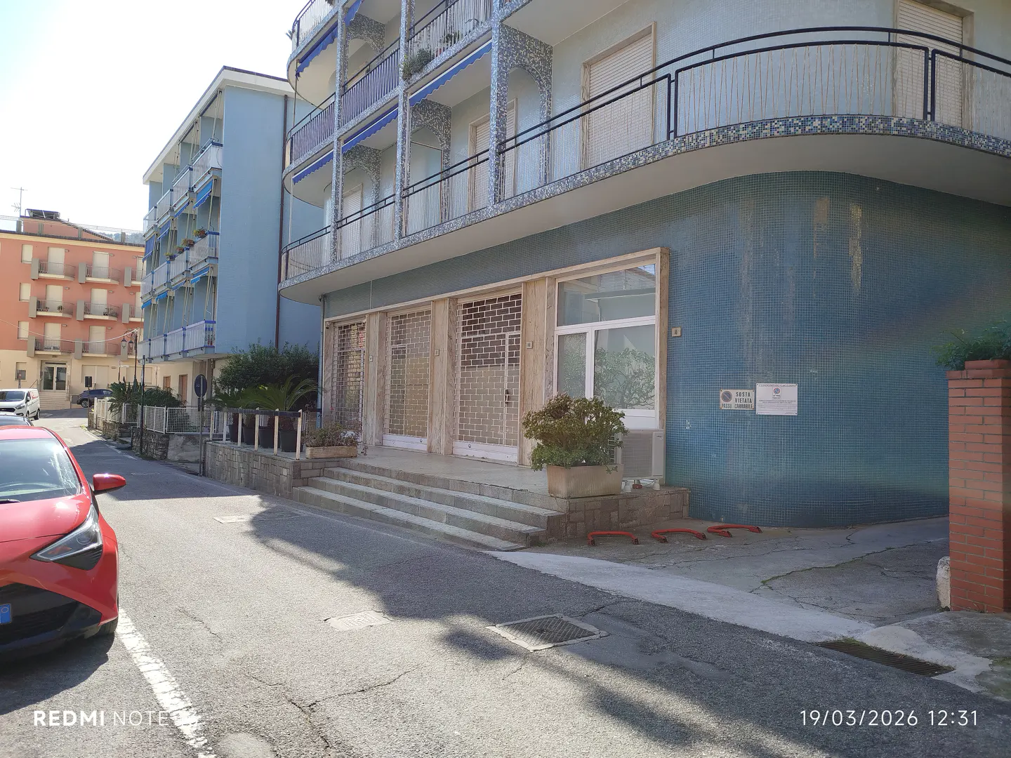 Exterior view of a blue building with balconies, metal security doors, and steps leading to the entrance. A red car is parked on the street.