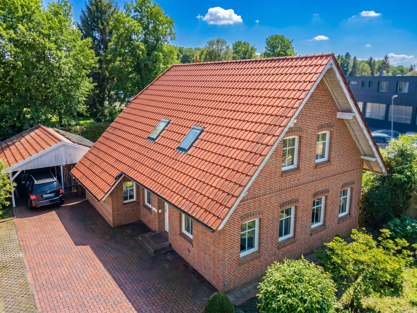 Aerial view of a two-story brick house with a red tile roof and white trim, a car port with a black car, and a brick driveway.