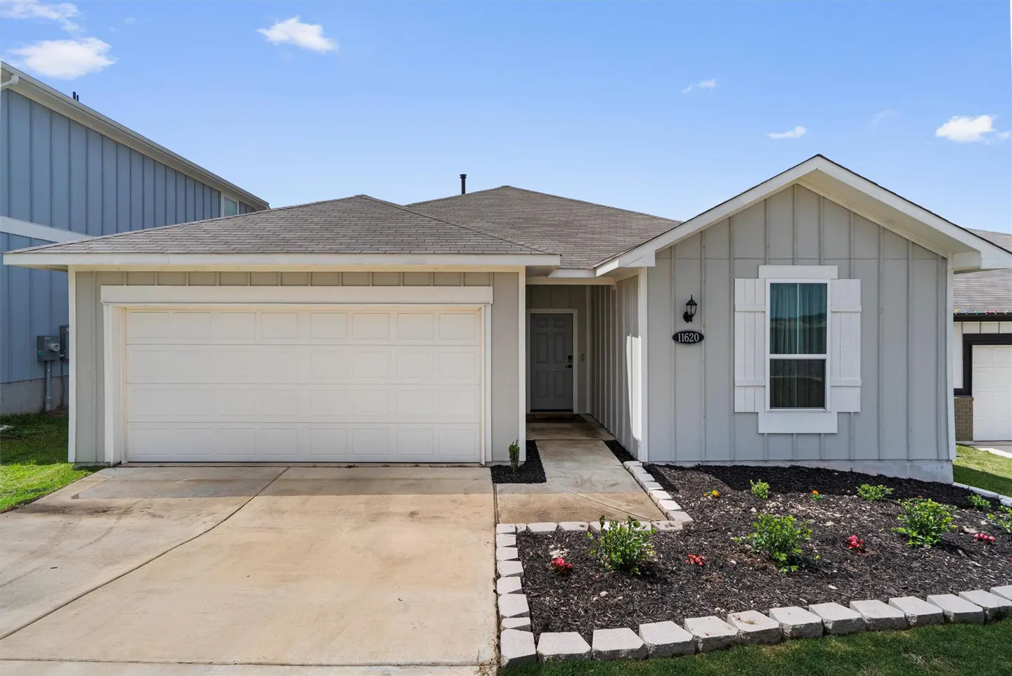 A single-story, light gray house with a white garage door, gray roof, and a small garden in front.