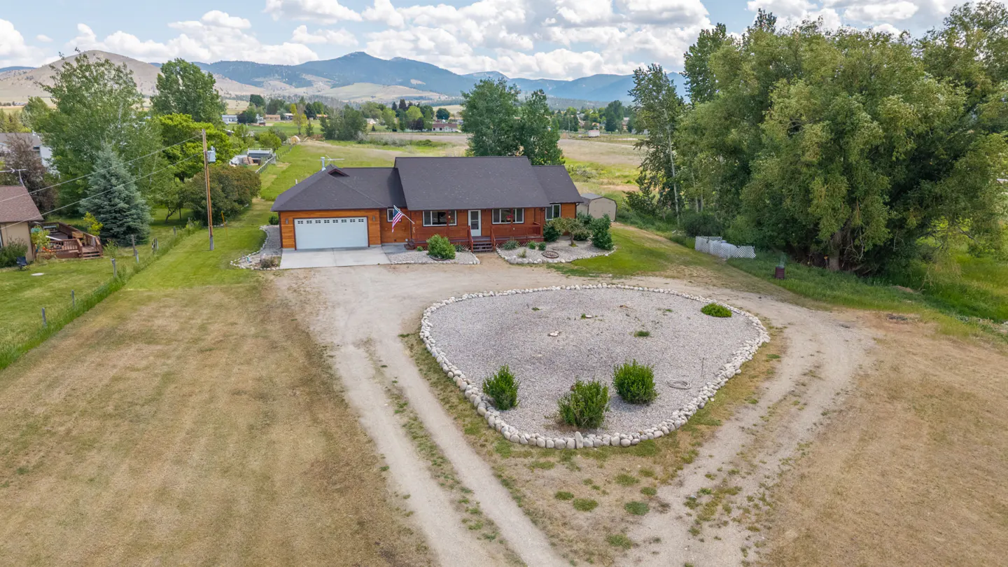 Aerial view of a brown, single-story house with a white garage door and a gravel driveway. Mountains and trees are in the background.