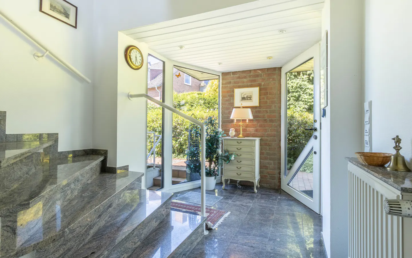 Bright entryway with polished grey stone stairs, a brick accent wall, a small chest of drawers, and a glass garden door.
