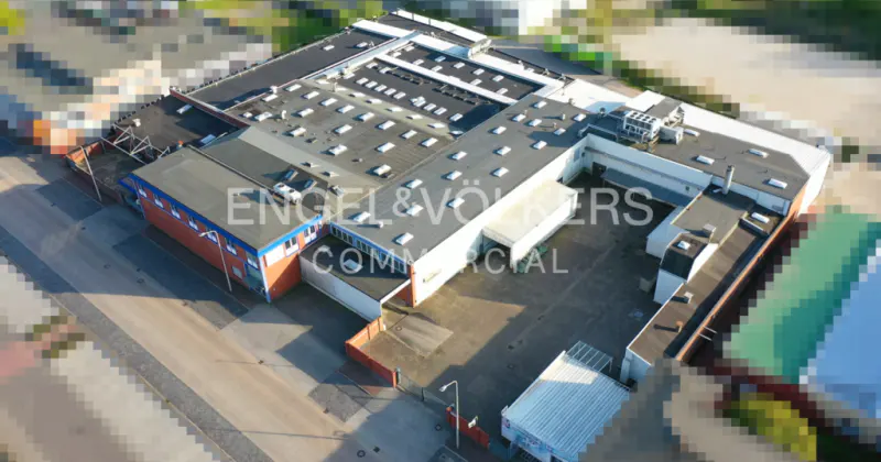 Aerial view of a large, flat-roofed commercial building with many skylights, surrounded by pavement and some greenery.