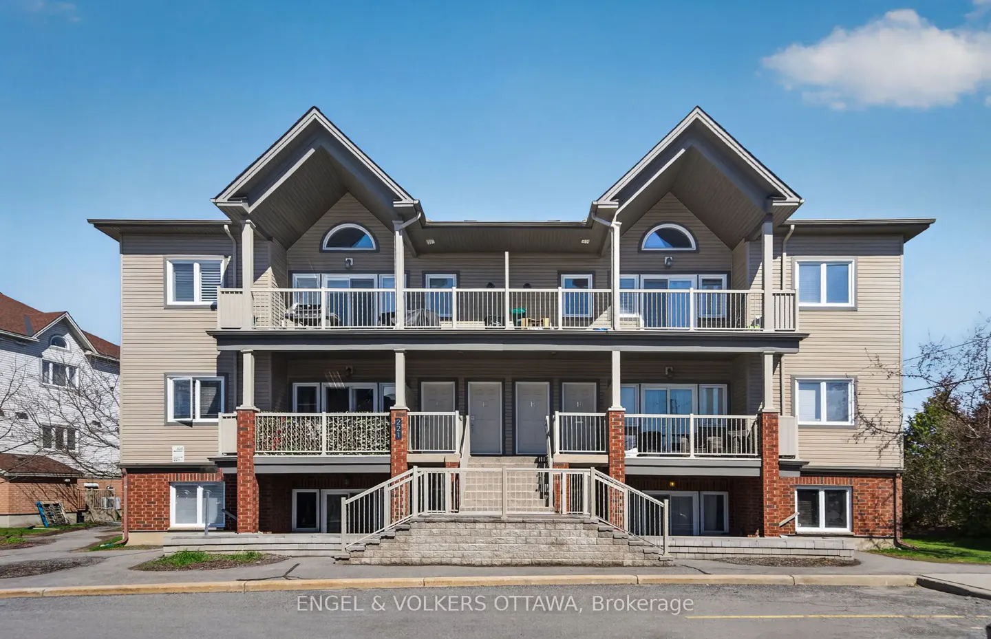 Two-story tan townhouse with white railings and brick accents under a blue sky.