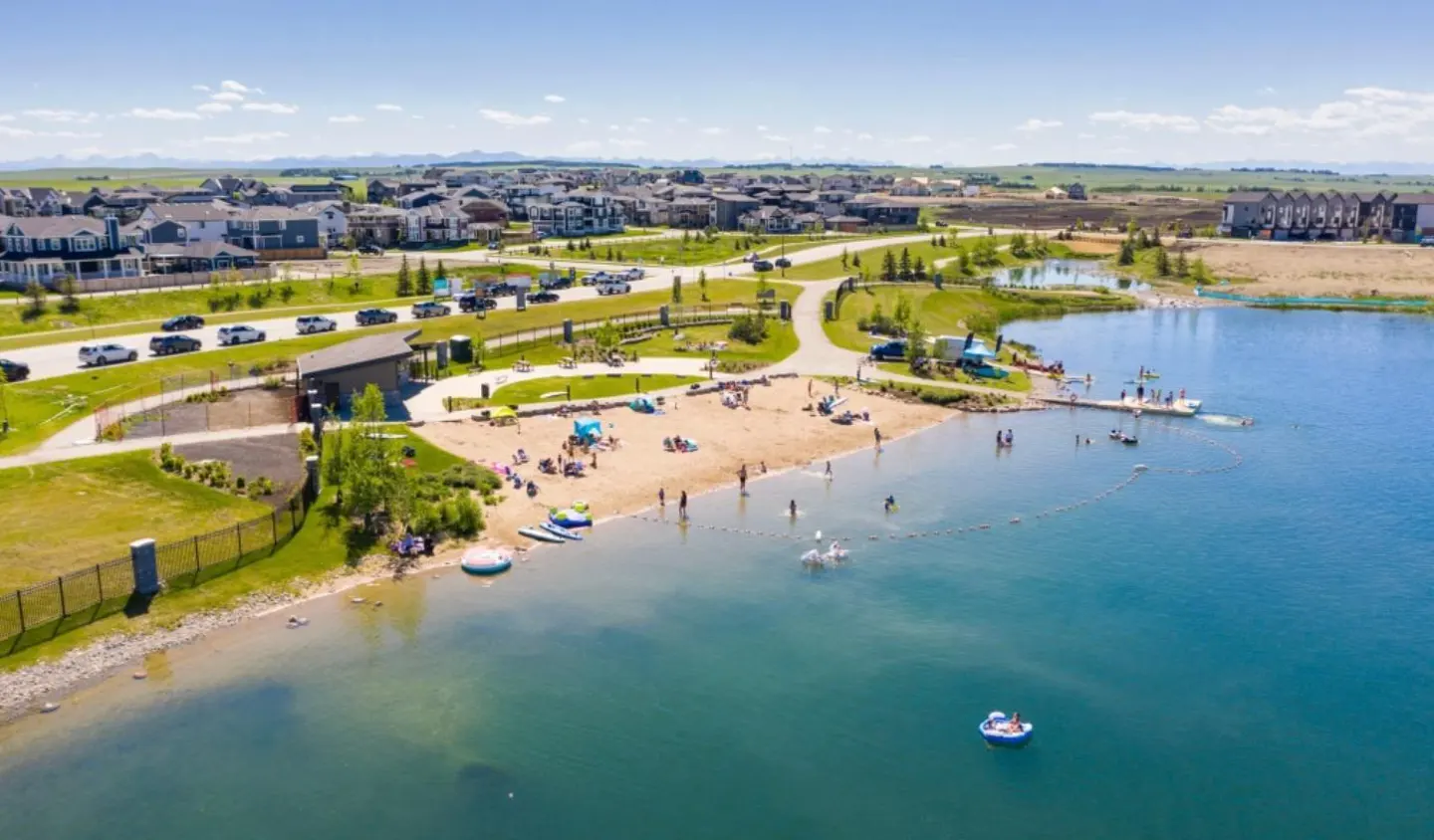 Aerial view of a community lake with a sandy beach, people swimming, and houses in the background on a sunny day.